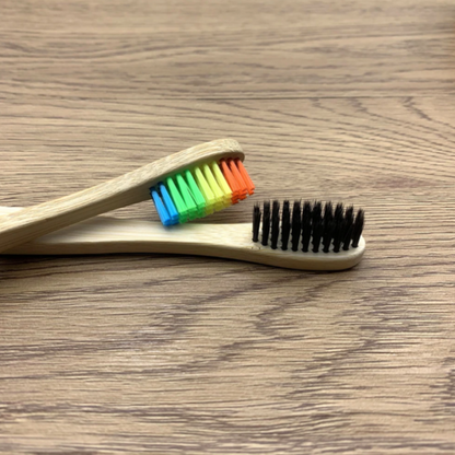 Two bamboo toothbrushes on a wooden surface, one with rainbow bristles and the other with black bristles.