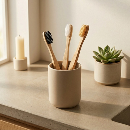 Three bamboo toothbrushes in a ceramic cup on a bathroom counter with a candle and plant in the background.
