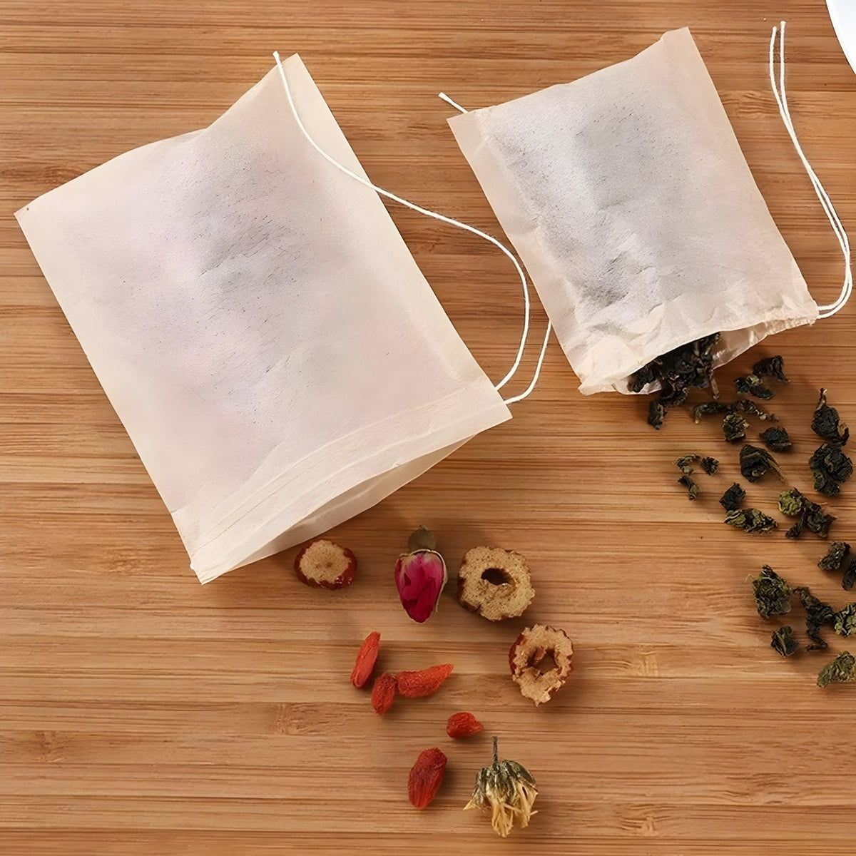 Two biodegradable drawstring tea bags on a bamboo wood surface, one open and spilling dried goji berries, rose buds and rolled oolong tea leaves.