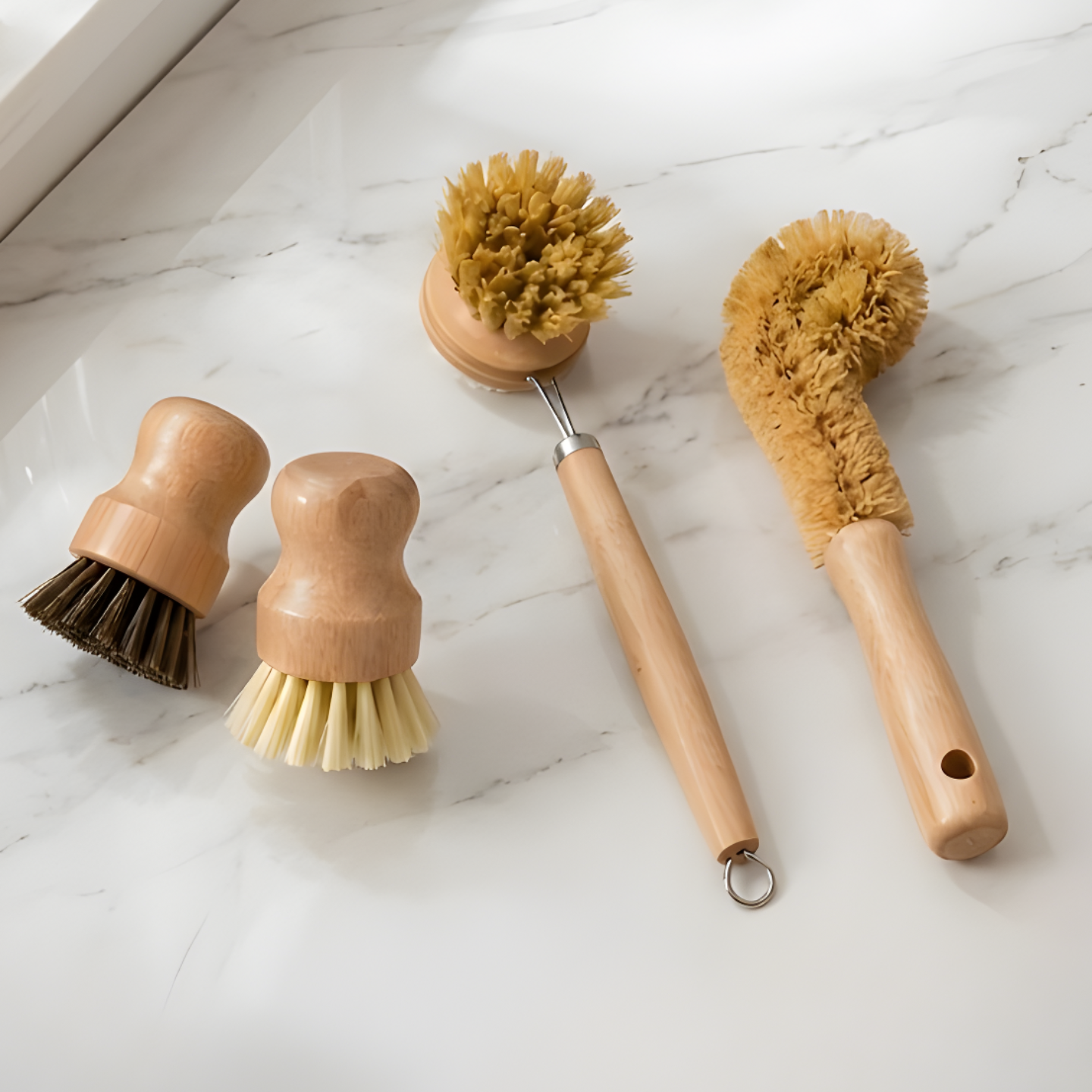 Four-piece natural sisal and wood kitchen brush set laid out on a white marble surface including two short-handled pot brushes, one long-handled bottle brush and one long-handled dish brush.