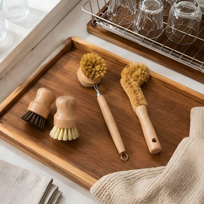Four natural wood and sisal cleaning brushes arranged on a dark wooden tray beside a dish rack and linen cloth in a kitchen setting