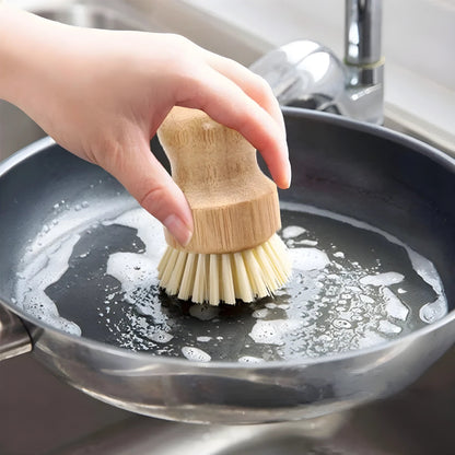 Hand scrubbing a non-stick frying pan with a short-handled natural wood and sisal pot brush under running water at a kitchen sink.