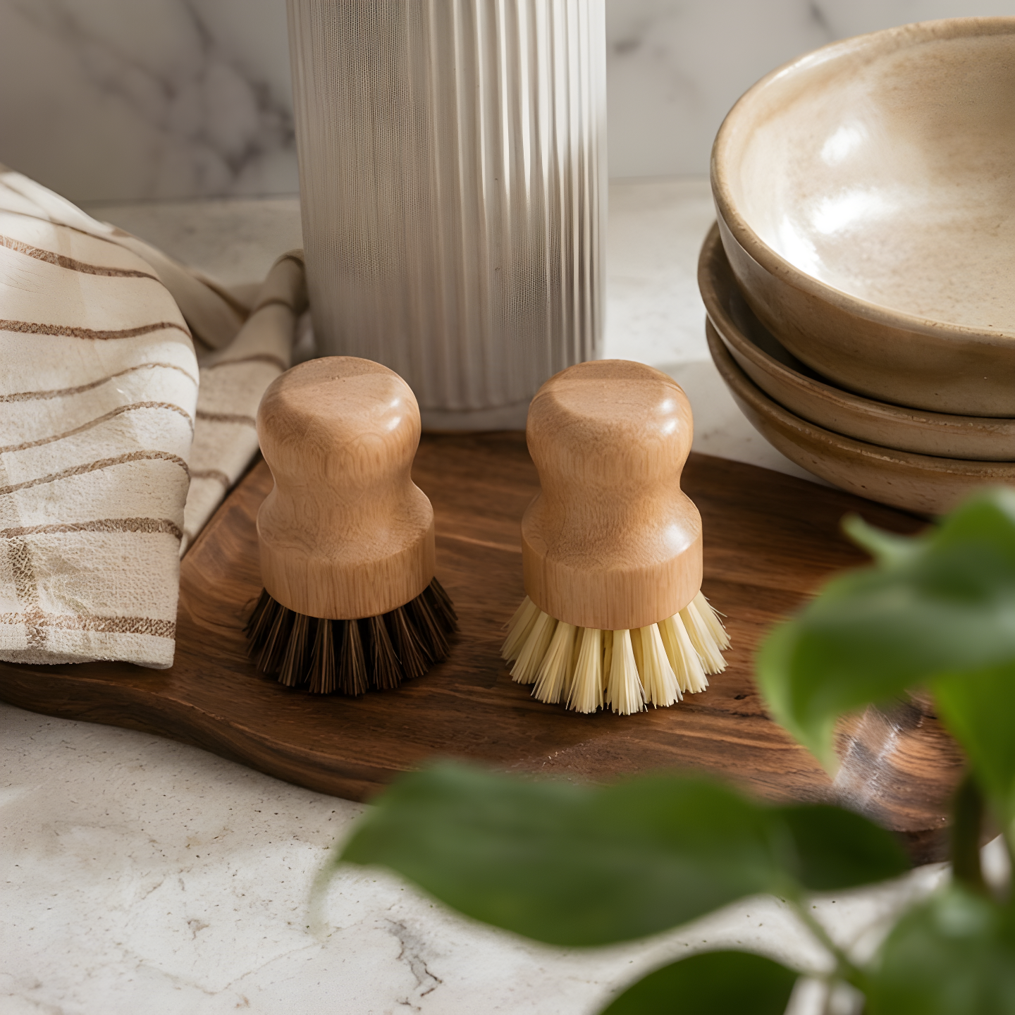  Two short-handled natural wood pot brushes with sisal and palm fibre bristles displayed on a wooden board beside stacked ceramic bowls and a green plant