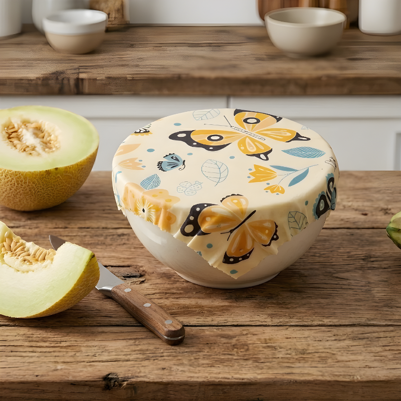 Reusable beeswax food wrap with butterfly print covering a bowl of melon on a rustic wooden kitchen counter, natural eco-friendly food storage keeping fruit fresh.
