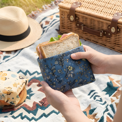 Hand holding a floral-patterned beeswax food wrap beside a wrapped sandwich on a picnic blanket, showing eco-friendly plastic-free food storage for outdoor use.