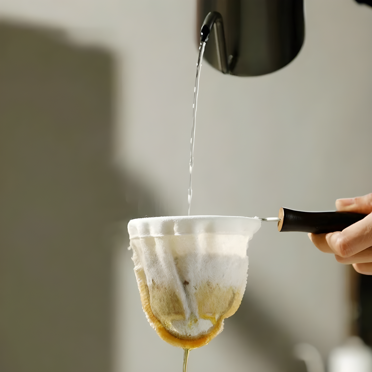 Close-up of coffee dripping from a reusable cloth filter into a cup, with steam rising and natural coffee oils visible on the cloth.