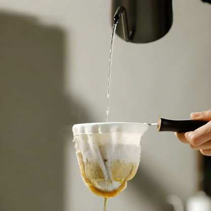 Close-up of coffee dripping from a reusable cloth filter into a cup, with steam rising and natural coffee oils visible on the cloth.