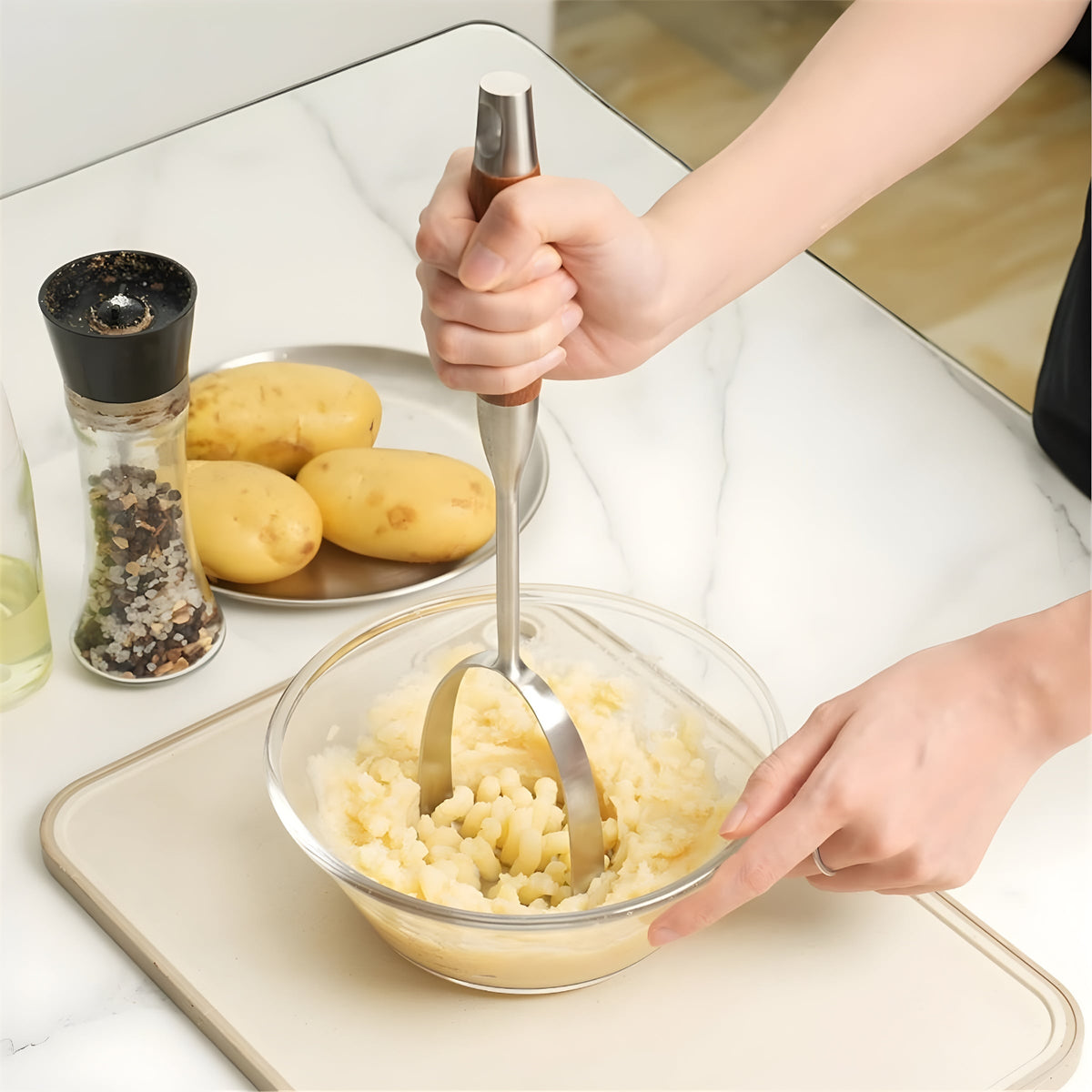 Hand mashing potatoes in a glass bowl with a rosewood handle masher on a marble counter