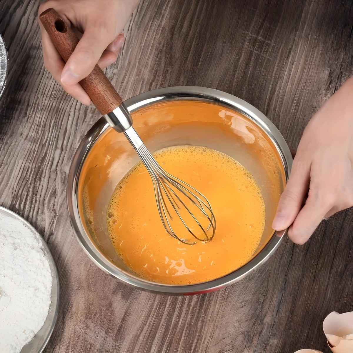 Hardwood handle whisk mixing batter in a stainless steel bowl on a wooden table