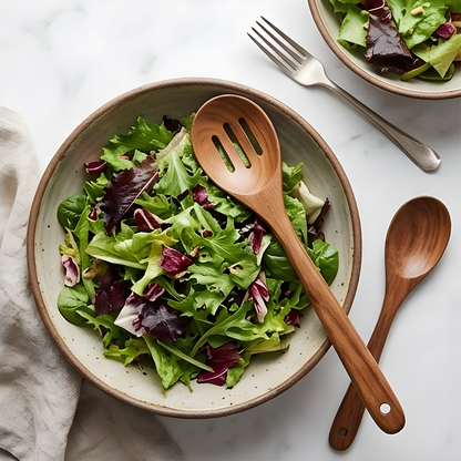 Teak salad fork and serving spoon resting in a ceramic bowl with fresh mixed green salad on a marble surface