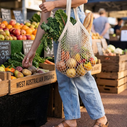 Woman shopping at an organic farmers market carrying a natural cotton mesh tote bag full of fresh vegetables and bread, reusable plastic-free shopping bag in real everyday use.